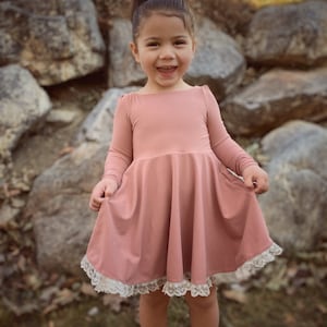 May include: A young girl wearing a dusty rose-colored dress with a white lace trim. The dress has long sleeves and a flared skirt. The girl is smiling and looking at the camera.