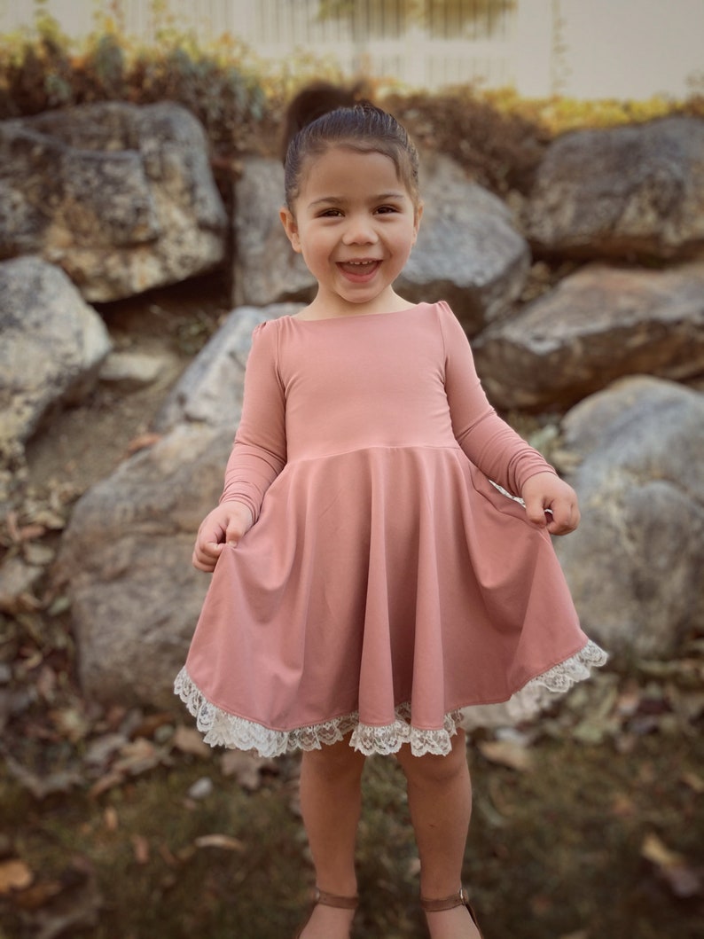May include: A young girl wearing a dusty rose-colored dress with a white lace trim. The dress has long sleeves and a flared skirt. The girl is smiling and looking at the camera.