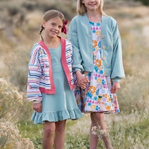 May include: Two girls wearing light blue dresses and cardigans. The girl on the left is wearing a striped cardigan with a pink trim. The girl on the right is wearing a solid cardigan with a floral print dress. Both girls are holding hands and smiling.