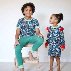 May include: Two children wearing matching pajamas with a Christmas dinosaur print. The boy is sitting on a wooden stool and the girl is standing. Both are wearing green pants and a blue shirt with green and red accents.