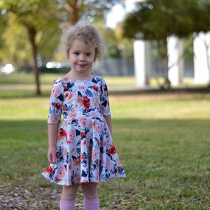 May include: A young girl with curly blonde hair wearing a white dress with a floral print. The dress has a round neckline and 3/4 sleeves. The girl is standing in a grassy area with trees in the background.