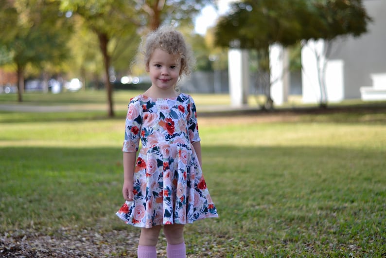 May include: A young girl with curly blonde hair wearing a white dress with a floral print. The dress has a round neckline and 3/4 sleeves. The girl is standing in a grassy area with trees in the background.