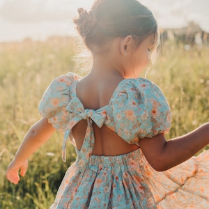 May include: A young girl wearing a light blue floral dress with a bow at the back. The dress has a tiered skirt and puffed sleeves. The girl is standing in a field of long grass.