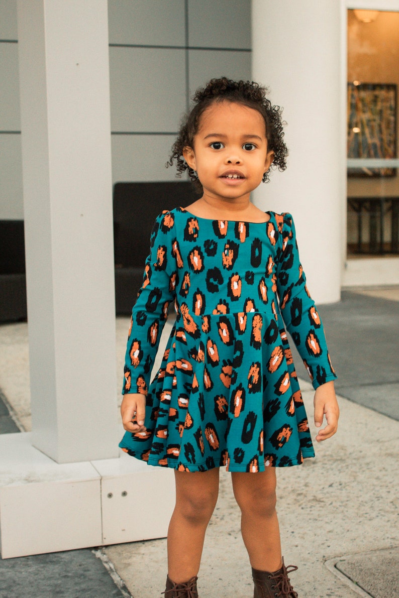 May include: A young girl wearing a teal blue dress with a brown and black leopard print. The dress has long sleeves and a flared skirt.