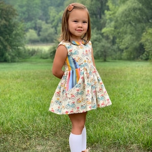 May include: A young girl wearing a white dress with a colorful bicycle print and white knee-high socks. She is standing in a grassy field with trees in the background.