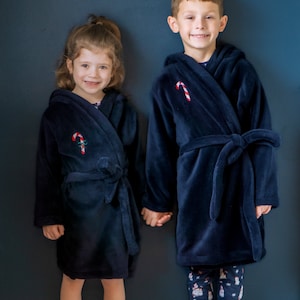 Może przedstawiać: Dwoje dzieci w dopasowanych, granatowych szlafrokach z haftem w kształcie cukierka. Dziewczynka stoi po lewej stronie, a chłopiec po prawej. Oboje się uśmiechają i trzymają się za ręce.