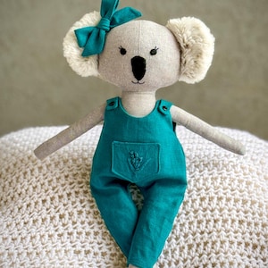 Peut inclure: Un jouet en peluche koala gris portant une salopette bleu sarcelle avec une broderie florale sur la poche. Le koala a un nez et des yeux noirs et un nœud bleu sarcelle sur la tête. Le jouet est assis sur une couverture en tricot blanc.