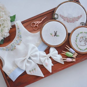 以下が含まれることがあります： 花柄の刺繍が施された白いノースリーブトップス、白いリボン2つ、花柄とウサギのデザインの刺繍枠3つ。枠は木製のトレイに糸巻きと一緒に置かれています。