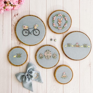 May include: Six embroidered hoops with various spring-themed designs. The hoops feature a bicycle with baskets of flowers, a bunny rabbit with flowers, a basket of eggs, a bird with flowers, and a basket of flowers. The hoops are all on a white wooden background.