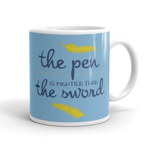 May include: A light blue ceramic mug featuring the dark blue text "the pen is mightier than the sword" centered on the front. Two yellow feather illustrations are positioned above and below the text, with a curved handle on the right side.