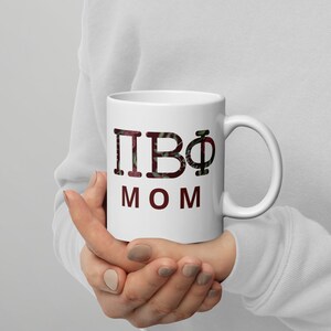 May include: White ceramic mug with a black and red design. The mug has the letters "ПВФ" and the word "MOM" printed on it.