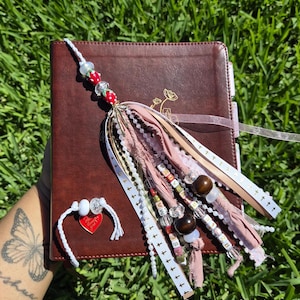 May include: A brown leather journal with a decorative tassel and a matching bracelet. The tassel features beads, ribbons, and charms in shades of pink, white, and brown. The bracelet has a red heart charm.