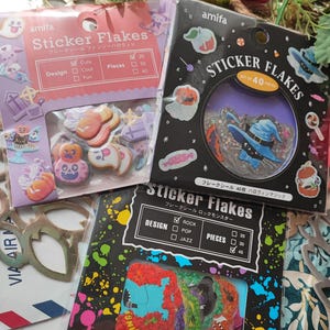 Puede incluir: Tres paquetes de copos de pegatinas con temática de Halloween. Cada paquete tiene un diseño y un esquema de color diferente. El primer paquete es rosa y morado con un fantasma y un sombrero de bruja. El segundo paquete es negro con un sombrero de bruja y una calabaza. El tercer paquete es negro y azul con un monstruo y una calavera.