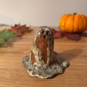 May include: A ceramic ghost ornament with a mottled brown, tan, and orange finish. The ghost has three eye holes and a wide base. An orange pumpkin and autumn leaves are in the background, creating a Halloween atmosphere.