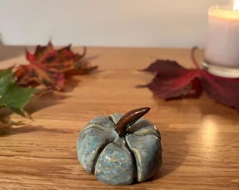 May include: A small, ceramic pumpkin with a teal and brown glaze. The pumpkin has a brown stem and is on a wooden surface. Autumn leaves and a lit candle are in the background.