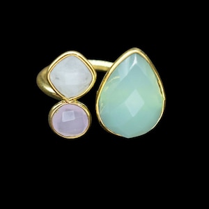 Pode incluir: Um anel dourado com três pedras preciosas: uma lágrima azul clara, um quadrado branco e um círculo rosa. As pedras são engastadas em biséis dourados sobre um fundo preto.