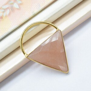 May include: A gold-toned triangle pendant with a pink gemstone. The pendant is on a white surface.