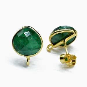 May include: A pair of emerald green gemstone stud earrings with a gold-colored setting. The earrings have a teardrop shape and are set on gold posts. The gemstones are faceted, reflecting light.