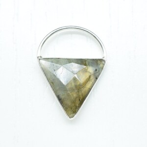 Puede incluir: Un colgante de plata con una piedra preciosa de labradorita triangular. El colgante tiene un diseño simple y minimalista.