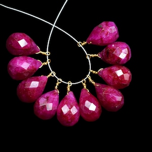 Puede incluir: Una hebra de cuentas de piedras preciosas de color rojo rubí, cada cuenta está facetada y tiene forma de lágrima. Las cuentas están enhebradas en un alambre fino con detalles dorados.