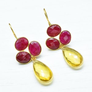 Könnte beinhalten: Goldene Ohrringe mit einem Cluster aus vier roten Edelsteinen und einem einzelnen tropfenförmigen gelben Edelstein.