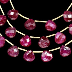 Pode incluir: Close-up de três fios de contas de rubi facetadas em forma de coração, enfiadas em fio dourado. Os rubis são de uma cor vermelha profunda e rica, e o fundo é preto.