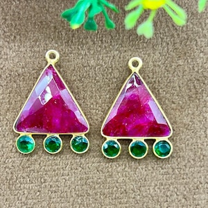 May include: Two gold-toned earrings with a triangular design. Each earring features a large, faceted, magenta-colored stone at the top and three small, round, green stones at the bottom. The earrings are displayed on a textured, tan surface.