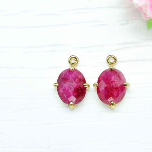 Op de afbeelding: Twee gouden ovale hangerbedels met een rode edelsteen in het midden. De bedels hebben een kraalbezetting en bevinden zich op een witte achtergrond.