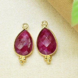 May include: Two gold-tone teardrop-shaped pendants with red gemstone accents. Each pendant has three small gold beads at the bottom.