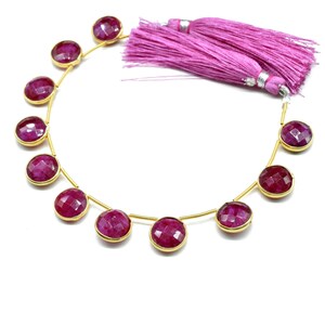 May include: A strand of ruby red gemstone beads with gold accents. The beads are round and faceted. The strand is strung on a gold wire and has a pink tassel at the end.