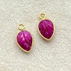 Peut inclure: Deux pendentifs en forme de feuille magenta vif, avec une texture sculptée et des bordures dorées. Chaque pendentif est doté d'une petite boucle pour être suspendu. Présentés sur une surface claire, ces bijoux sont élégants.