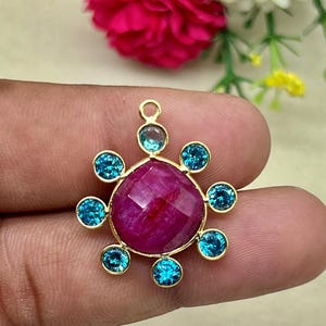 May include: A gold-toned pendant featuring a large, faceted, deep pink gemstone at its center, surrounded by eight smaller, round, light blue gemstones. The pendant has a loop at the top for a chain.