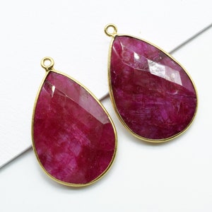 Op de afbeelding: Twee traanvormige hangers van robijn edelstenen met goudkleurige bezels en springringen. De edelstenen zijn diep rood van kleur en hebben een gefacetteerde afwerking.