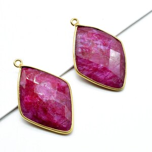 May include: Two gold-toned pendant charms with a diamond-shaped ruby gemstone. The ruby is a deep pink color with a faceted cut.