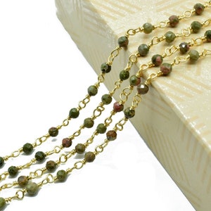 Op de afbeelding: Een gouden ketting met groene en rode edelsteen kralen. De ketting ligt op een beige oppervlak met een geometrisch patroon.