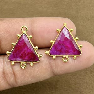 May include: A pair of gold-colored earrings with triangular, magenta-colored gemstones. The earrings have a gold frame with small, round accents and a loop at the top and bottom for attachment.