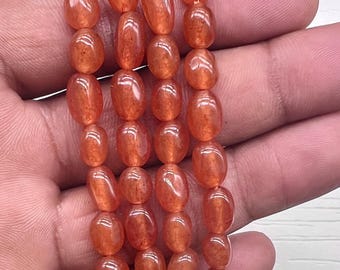 Puede incluir: Hebras de cuentas ovaladas de color naranja rojizo. Las cuentas están ensartadas, con ensartado visible en la parte inferior. Las cuentas tienen un acabado brillante y se sostienen en una mano sobre un fondo blanco.