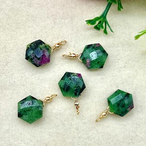 Puede incluir: Cinco pendientes hexagonales facetados con un lazo de alambre dorado. Los pendientes son de color verde oscuro con motas rojas y moradas. Los pendientes se exhiben sobre una superficie de color crema, con flores rosas y blancas en el fondo.