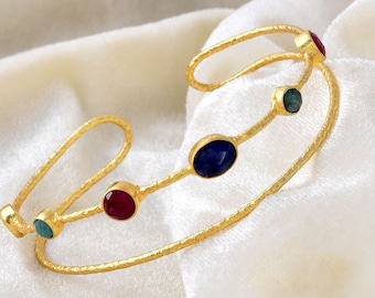 May include: Gold bangle bracelet with a textured finish and four oval-shaped gemstones. The gemstones are blue, green, and red.
