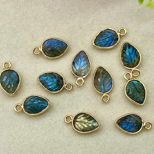 May include: A collection of teardrop-shaped pendants with a leaf design. Each pendant features a gold-colored bezel and a shimmering blue and green stone. The pendants are arranged on a light-colored surface.