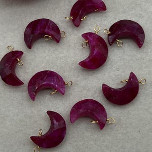 May include: A collection of deep magenta crescent moon-shaped gemstone pendants. Each pendant has a gold-colored wire loop for stringing. The gemstones have a faceted surface and are arranged on a light gray background.