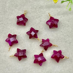 Puede incluir: Primer plano de varias gemas facetadas en forma de estrella, de un tono magenta intenso. Cada estrella está cableada individualmente con alambre dorado, algunas conectadas en cadena. Las gemas están dispuestas sobre una superficie clara.