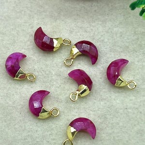 Puede incluir: Primer plano de varios dijes en forma de media luna. Cada dije presenta una piedra preciosa facetada, de color magenta intenso, con una tapa y un lazo de metal dorado. Los dijes están dispersos sobre una superficie gris claro, lo que sugiere componentes de joyería.