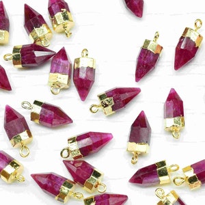 Może przedstawiać: Kolekcja zawieszek z rubinowych kamieni szlachetnych z pozłacanymi akcentami. Każda zawieszka ma fasetowany, spiczasty kamień szlachetny z pozłacaną pętelką u góry do przymocowania do biżuterii.