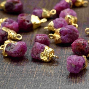 May include: A collection of pink ruby gemstone pendants with gold-plated accents. The raw, natural ruby stones are irregular in shape and have a rough, textured surface. Each pendant has a gold loop for attaching to jewelry.