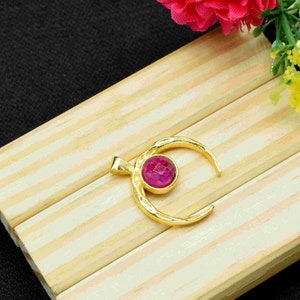 May include: A gold crescent moon pendant with a pink gemstone in the center. The pendant is on a wooden surface.