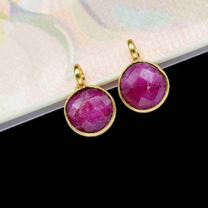 May include: Two gold-toned charms with pink gemstone accents. The charms are round and have a faceted design.