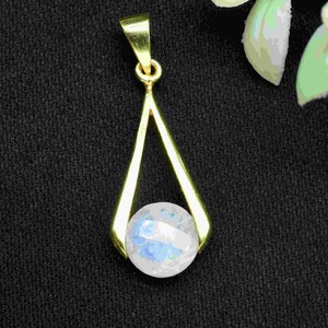 May include: A gold-colored pendant with a teardrop design, featuring a faceted, iridescent, round gemstone. The pendant hangs from a small loop. The background is a dark, textured fabric, with a few green leaves and white petals in the upper right corner.