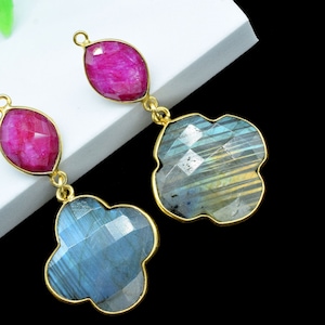 以下が含まれることがあります： ピンクとブルーの宝石のドロップが付いたゴールドのイヤリングのペア。イヤリングは、クローバー型のブルーの宝石と、ピンクのオーバル型の宝石が特徴です。