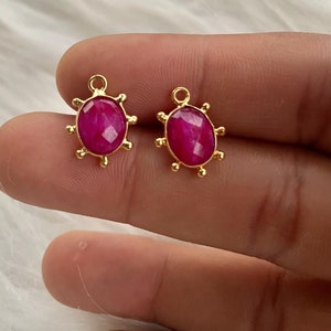 May include: A pair of gold-toned earrings with pink oval-shaped gemstones. The earrings have a decorative design around the gemstones.